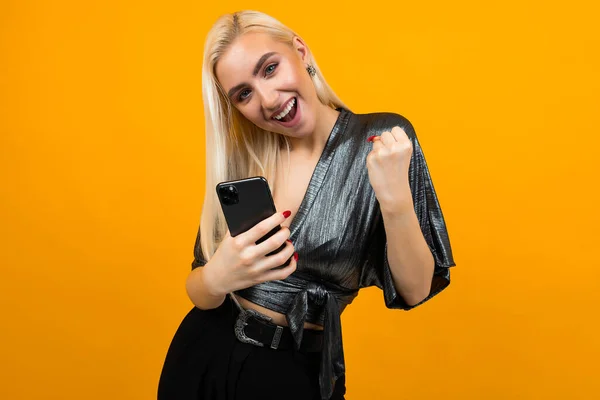 Happy charming young woman smiles with joy holding a phone in her hands on a yellow studio background — Stock Photo, Image