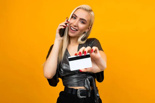 Menina alegre faz compras por telefone segurando um cartão de crédito com um mockup em um fundo de estúdio amarelo — Fotografia de Stock