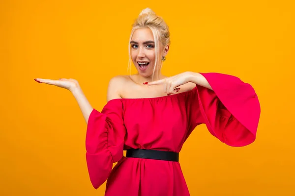 Encantadora chica rubia sonriente en un vestido rojo con una mano para la publicidad de cosméticos en un fondo de estudio amarillo — Foto de Stock