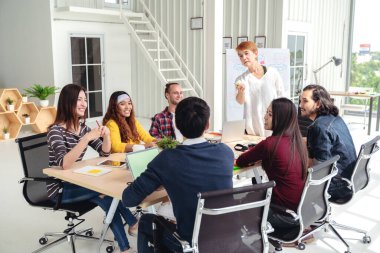 Modern ofis konseptinde toplanan, çok ırklı, yaratıcı, gülen ve beyin fırtınası yapan bir grup genç. Diğerleri yan yana otururken, kadınlar paylaşmak için ayakta duran el hareketi yapıyorlar..