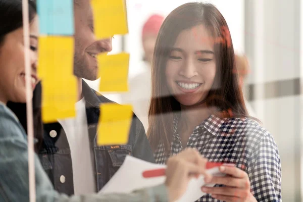 Bir grup başarılı yaratıcı, etnik çeşitlilikteki takım gülüşü ve modern ofiste nota veya yapışkan notla proje üzerine beyin fırtınası yapıyor. Beyaz adam cam duvar paylaşımı fikrini işaret ediyor..