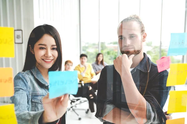 Group of young successful creative multiethnic team smile and brainstorm on project together in modern office. Woman sticking and look at sticky note on glass wall sharing idea with engaged cheerful.