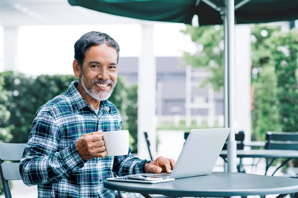 Mutlu beyaz kısa sakallı, olgun bir adamın portresi. Laptop kullanırken kahve içiyor. Emekli İspanyol asıllı, yetişkin Asyalı bir adamın modern kafede oturup gülümsemesi sıradan bir şehir hayatı..