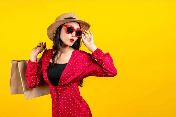 Asian trendy shopaholic woman excited about new purchases or sales holding shopping bags and looking to side copy space over yellow background. Happy beautiful Asian woman carrying shopping bags.