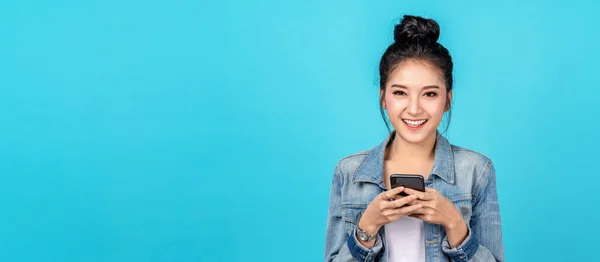 Portre Sancağı mutlu Asyalı kadın mutluluğu hissediyor ve kameraya bakıyor mavi arka planda akıllı telefonu tutuyor. Hoş Asyalı kız günlük kot gömlek giyerek gülümsüyor ve internet üzerinden alışveriş yapıyor.