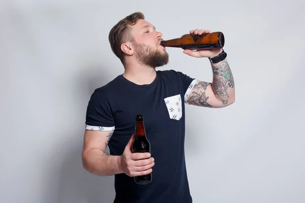 Brutální vousatý muž s Tetovaný ruku pije pivo z láhve. — Stock fotografie