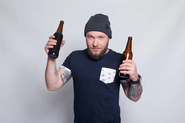 Homem barbudo brutal com braço tatuado bebe uma cerveja de uma garrafa . — Fotografia de Stock