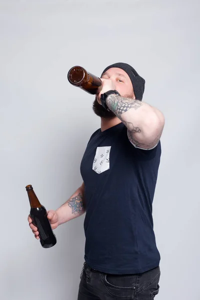 Brutal bearded male with tattooed arm drinks a beer from a bottle.