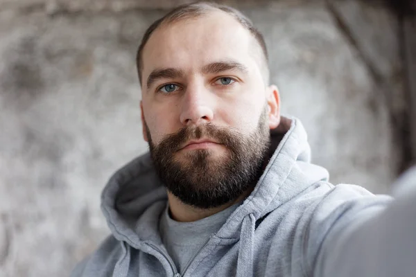 Homem barbudo em um fundo cinza faz um selfi — Fotografia de Stock