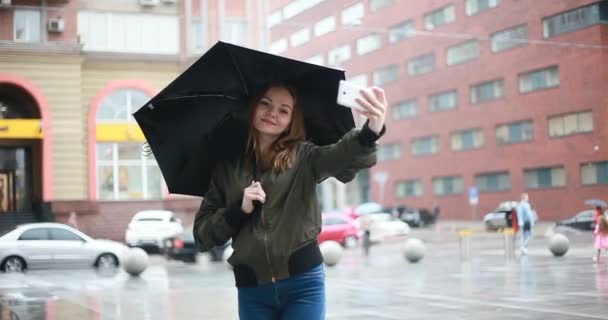 の概念 若いブルネットの女性は雨の日に傘手敷地内に立っています 女の話が電話では Selfie メッセージ 女性が携帯電話を見ての書き込み — ストック動画