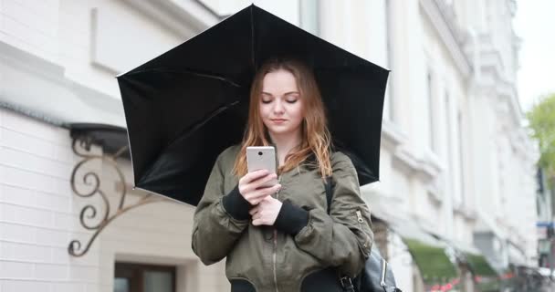 の概念 若いブルネットの女性は雨の日に傘手敷地内に立っています 女の話が電話では Selfie メッセージ 女性が携帯電話を見ての書き込み — ストック動画