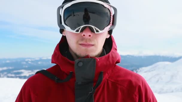 ポートレートクローズアップクールなハンサムな男若いスノーボーダー外冬のスキーリゾート探しカメラ陽気な笑顔青い目ヘルメットアクティビティスポーツライフスタイル極端な男性の山スキー人 — ストック動画