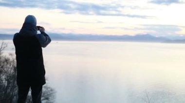 Adam akıllı telefonuyla Sunset 'te karlı vahşi bir arazide fotoğraf çekiyor. Yalnız bir yetişkinin arka planda kalan vahşi deniz manzaralı güzel resimlerine bakması..
