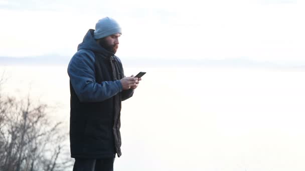 男は夕日の雪の野生の風景の中に彼のスマートフォンで写真を撮ります 一人の孤独な大人の男が行くことによって時間を費やしているの背景と野生の海辺のエリアの美しさの写真を見て回る — ストック動画