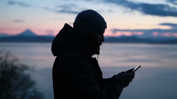 在日落时 人类带着他的智能手机在雪地的野景中拍照 回头看一个孤独的成年人 他正呆在一边 环顾四周 欣赏着野外海滨的美景 — 图库视频影像
