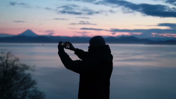 男は夕日の雪の野生の風景の中に彼のスマートフォンで写真を撮ります 一人の孤独な大人の男が行くことによって時間を費やしているの背景と野生の海辺のエリアの美しさの写真を見て回る — ストック動画