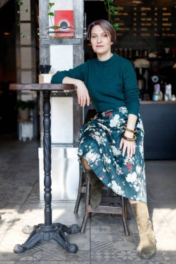 Kafede kahve içerken gülümseyen kameraya bakan güzel bir kız. Yeşil örülmüş süveter. Şık giyimli kız. Kız telefona bakıyor. İş kadını