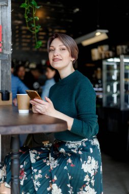 Kafede kahve içerken gülümseyen kameraya bakan güzel bir kız. Yeşil örülmüş süveter. Şık giyimli kız. Kız telefona bakıyor. İş kadını