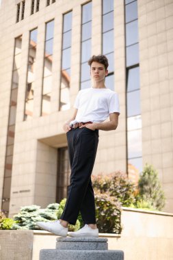 Modaya uygun siyah tişörtlü siyah tişörtlü, siyah pantolonlu, modaya uygun saç stili modern bir iş merkezinin yanında duruyor. Bir yaz gününde sokakta çekici bir adam..