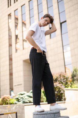 Modaya uygun siyah tişörtlü siyah tişörtlü, siyah pantolonlu, modaya uygun saç stili modern bir iş merkezinin yanında duruyor. Bir yaz gününde sokakta çekici bir adam..