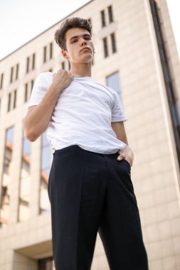 Modaya uygun siyah tişörtlü siyah tişörtlü, siyah pantolonlu, modaya uygun saç stili modern bir iş merkezinin yanında duruyor. Bir yaz gününde sokakta çekici bir adam..