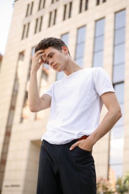 Modaya uygun siyah tişörtlü siyah tişörtlü, siyah pantolonlu, modaya uygun saç stili modern bir iş merkezinin yanında duruyor. Bir yaz gününde sokakta çekici bir adam..