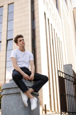 Modaya uygun siyah tişörtlü siyah tişörtlü, siyah pantolonlu, modaya uygun saç stili modern bir iş merkezinin yanında duruyor. Bir yaz gününde sokakta çekici bir adam..