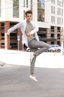 Beyaz gömlekli ve gri pantolonlu şık saç kesimi olan bir erkek model. Modayı takip eden saç stili modern bir iş merkezine yakın. Genç adam son moda bir kıyafetle atlıyor.