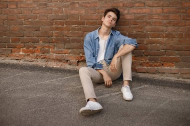 Genç hippi mavi kot gömleğiyle oturur ve etiketsiz beyaz pamuklu bir tişörtü tuğla duvar arkasında gösterir.