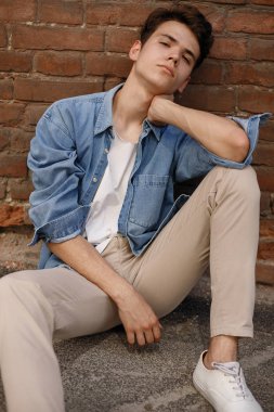 Genç hippi mavi kot gömleğiyle oturur ve etiketsiz beyaz pamuklu bir tişörtü tuğla duvar arkasında gösterir.