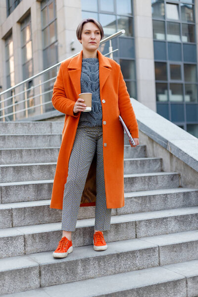 Стильная женщина в ярко-оранжевом пальто на городской сцене, используя ноутбук и работая над ним во время перерыва. Технология наружного освещения. пить кофе, чай, какао
