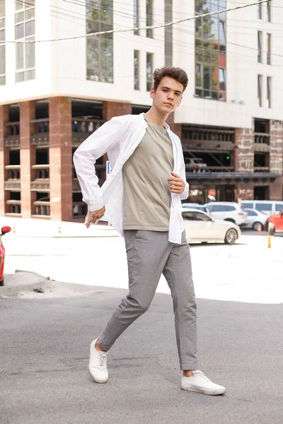 guy model with a stylish haircut posing outdoors in a white shirt and gray trousers. trendy hairstyle rests near a modern business center. Attractive guy in the street on a summer day.