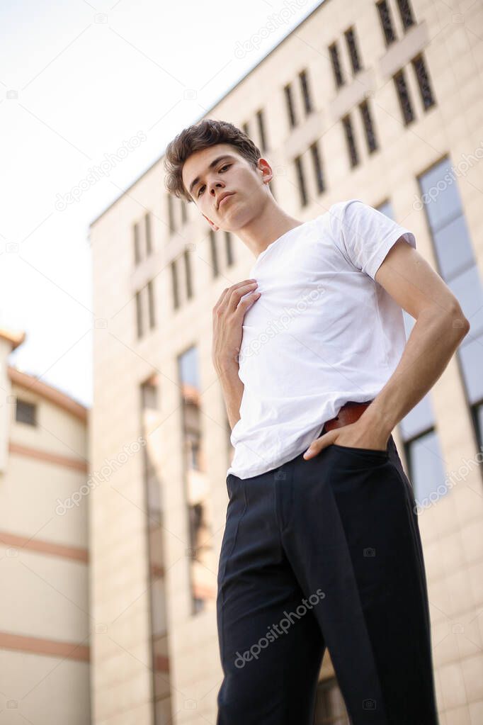 Elegant young man in a fashionable black shirt in a white stylish t-shirt in black pants with a trendy hairstyle rests near a modern business center. Attractive guy in the street on a summer day.