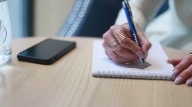 Bir kadının günlüğüne oturup bir şeyler yazmasına yakın çekim. Uzun saçlı sarışın kadın modern bir kafede oturuyor ve defterine bazı notlar yazıyor. Bir dizüstü bilgisayarın nişan alınmış görüntüleri