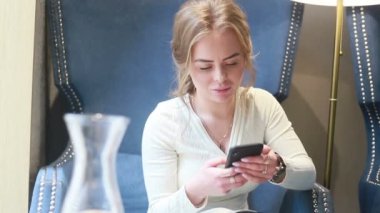 Kafeteryada telefonu kullanıp gülümseyen beyaz bir kadının portresi. Kaydır, hafifçe vur. Mesajlaşma, video izleme, blog ya da içerik. Yaklaşın, ağır çekim.