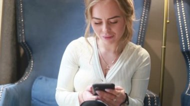 Kafeteryada telefonu kullanıp gülümseyen beyaz bir kadının portresi. Kaydır, hafifçe vur. Mesajlaşma, video izleme, blog ya da içerik. Yaklaşın, ağır çekim.