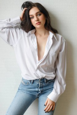 Açık gri arka planda çıplak omuzları olan kabarık bir gömlek giyen genç bir kadının portresi. Kot pantolonlu ve beyaz tişörtlü güzel esmer kız gülümsüyor.