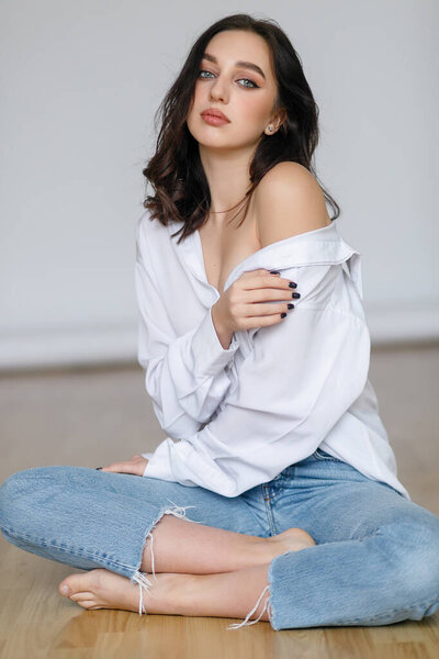 портрет сидящей молодой женщины в роскошной рубашке с голыми плечами на светло-сером фоне. Красивая брюнетка в джинсах и белой футболке улыбается
