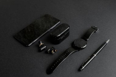 Tek renkli arka planda erkek güzelliği için aksesuarlar. Siyah kalem, siyah akıllı saat, akıllı telefon ve karanlık arka planda kablosuz kulaklık. Yukarıdan bak. Minimalist siyah eğilim 2020.