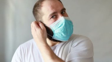 Koruyucu maskeli bir adam öksürüyor. Adam hasta, üşütmüş, öksürüyor. Beyaz koruyucu takım elbiseli beyaz sakallı adam kameraya baktı ve yüzüne tıbbi bir maske taktı. Kapat.