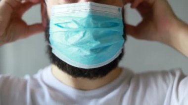 Koruyucu maskeli bir adam öksürüyor. Adam hasta, üşütmüş, öksürüyor. Beyaz koruyucu takım elbiseli beyaz sakallı adam kameraya baktı ve yüzüne tıbbi bir maske taktı. Kapat.