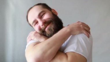 Yakışıklı bir adam mutlu ve olumlu bir şekilde kendine sarılıyor, kendinden emin gülümsüyor. Kendini sevme ve kendini koruma. Stüdyo videosu