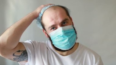Koruyucu maskeli bir adam öksürüyor. Adam hasta, üşütmüş, öksürüyor. Beyaz koruyucu takım elbiseli beyaz sakallı adam kameraya baktı ve yüzüne tıbbi bir maske taktı. Kapat.