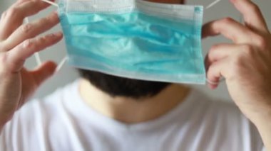 Koruyucu maskeli bir adam öksürüyor. Adam hasta, üşütmüş, öksürüyor. Beyaz koruyucu takım elbiseli beyaz sakallı adam kameraya baktı ve yüzüne tıbbi bir maske taktı. Kapat.