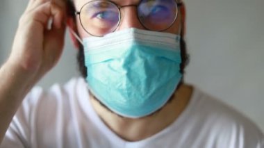 Koruyucu maskeli bir adam öksürüyor. Adam hasta, üşütmüş, öksürüyor. Beyaz koruyucu takım elbiseli beyaz sakallı adam kameraya baktı ve yüzüne tıbbi bir maske taktı. Kapat.