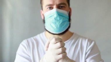 Koruyucu maskeli bir adam öksürüyor. Adam hasta, üşütmüş, öksürüyor. Beyaz koruyucu takım elbiseli beyaz sakallı adam kameraya baktı ve yüzüne tıbbi bir maske taktı. Kapat.