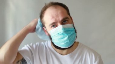 Koruyucu maskeli bir adam öksürüyor. Adam hasta, üşütmüş, öksürüyor. Beyaz koruyucu takım elbiseli beyaz sakallı adam kameraya baktı ve yüzüne tıbbi bir maske taktı. Kapat.