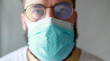 Koruyucu maskeli bir adam öksürüyor. Adam hasta, üşütmüş, öksürüyor. Beyaz koruyucu takım elbiseli beyaz sakallı adam kameraya baktı ve yüzüne tıbbi bir maske taktı. Kapat.