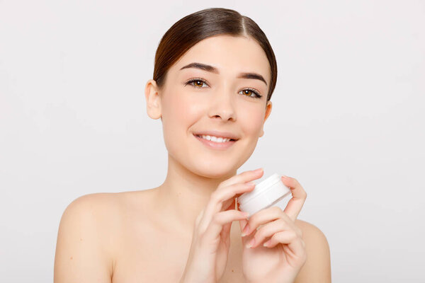 Beauty portrait of girl with perfect nude make-up. Holding cream, some cream on finger. Looking at camera and smiling. Beauty salon. Head and shoulders, studio, indoors