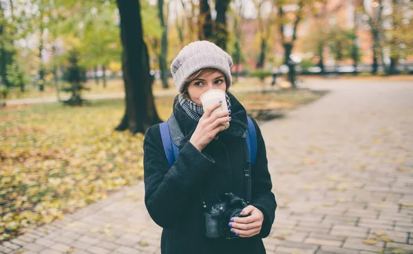 Frau im Mantel mit einer Tasse Kaffee-Latte mit Milch. einsame Frau steht auf einer verschneiten, herbstlich verlassenen Straße im Park. — Stockfoto
